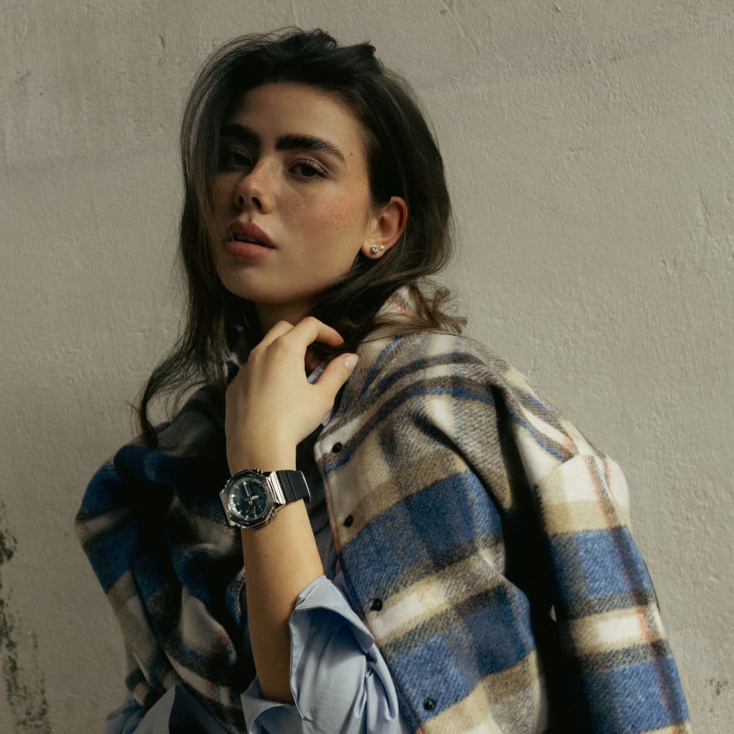 Un mannequin féminin regardant directement l'appareil photo et portant une veste à carreaux sans serrer les épaules. On la voit clairement porter la nouvelle montre GBM-2100 de G-SHOCK avec une lunette en métal.