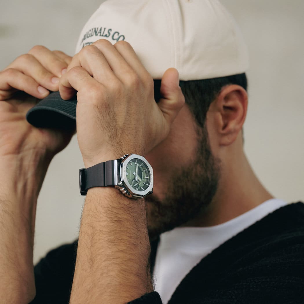 Le modèle tient sa casquette sur la tête, l'accent est mis sur son poignet : Il porte la montre GBM-2100A-1A3 de G-SHOCK
