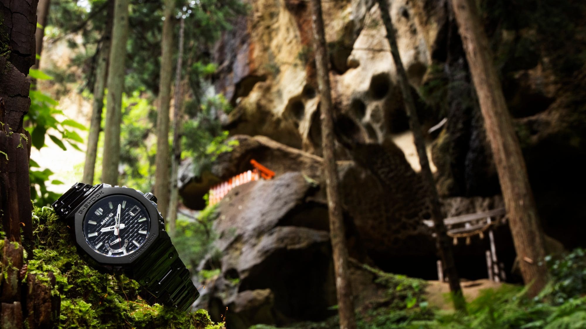La montre G-SHOCK MRG-B2100B, avec son bracelet noir et son boîtier noir, est posée dans une forêt sur une pierre recouverte de mousse. 
