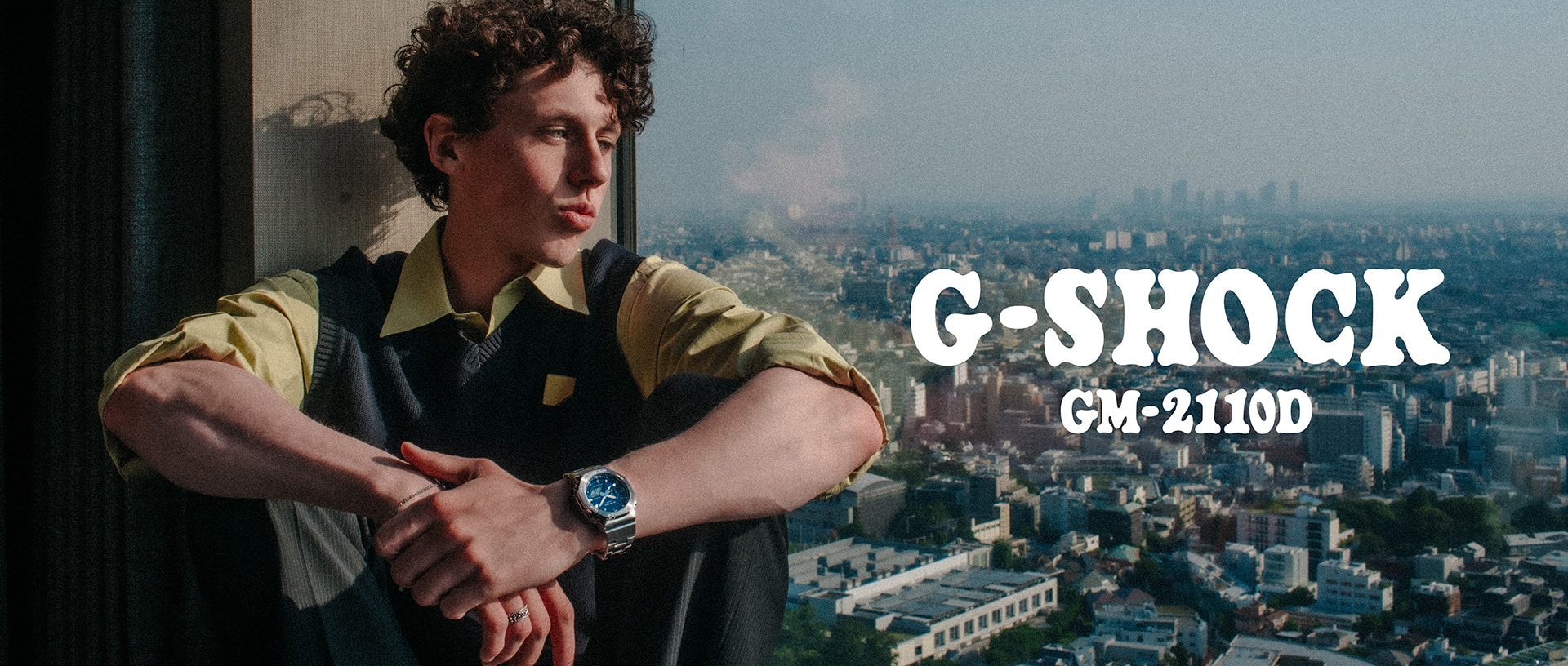 A man wearing a G-SHOCK GM-2110D watch sitting by a window.
