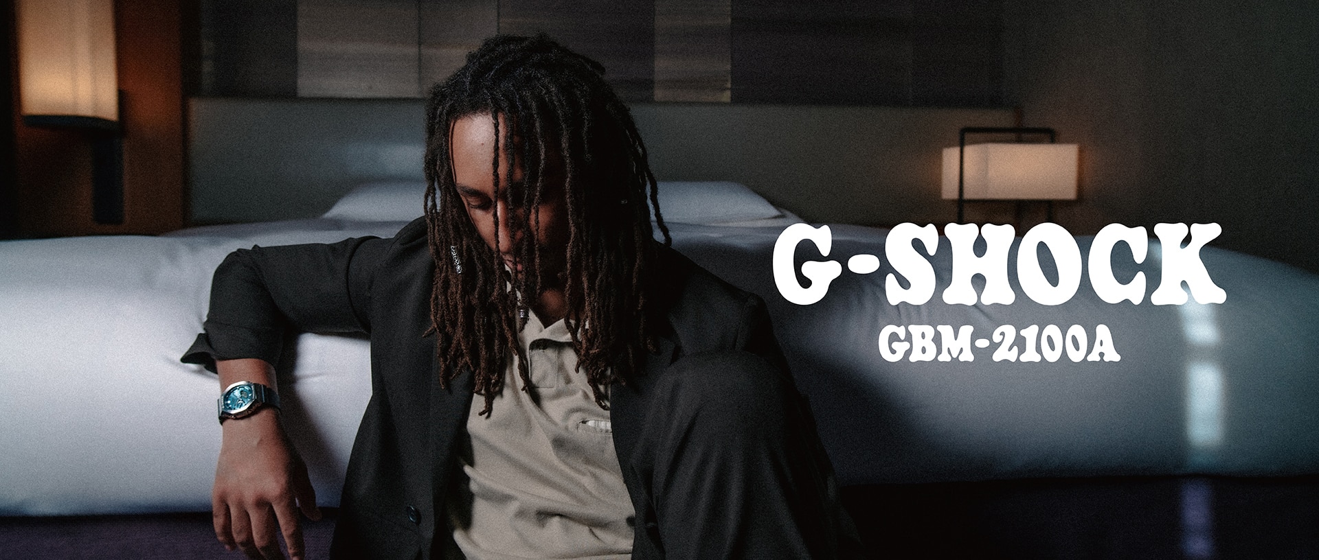 A man wearing a G-SHOCK GBM-2100A watch sitting near a bed.