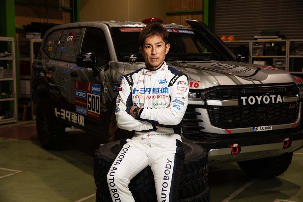 Miura Akira sits on the tire in front of the Land Cruiser, with his arms crossed.