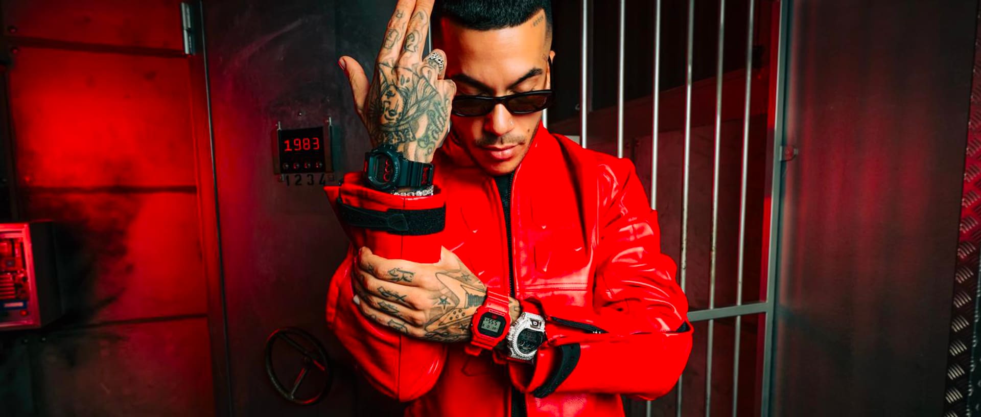 Il rapper Sfera Ebbasta, con indosso una giacca di pelle rossa, è in piedi davanti a una cassaforte circondata da una grata. Si afferra l'avambraccio, rivolto verso l'alto, e ai polsi indossa tre  orologi digitali G-SHOCK neri, bianchi e rossi.