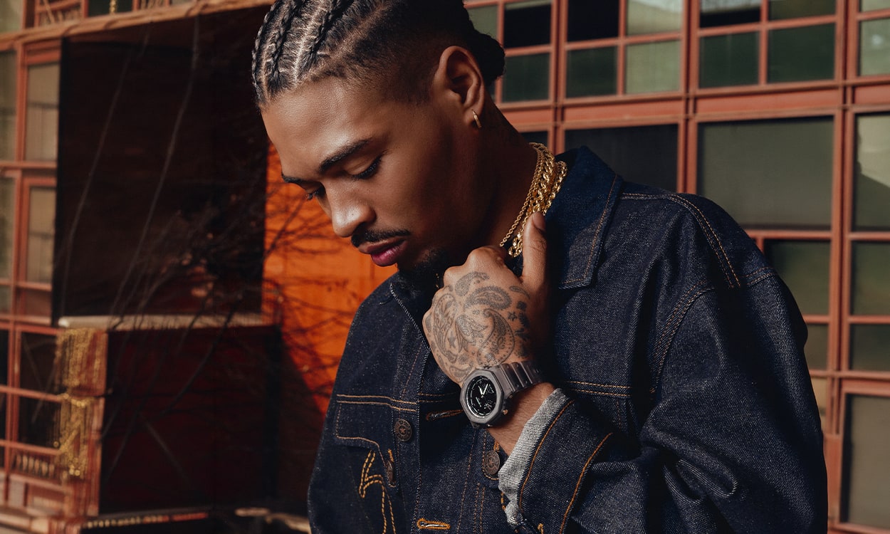 Artie 5ive in posa con una giacca di jeans scuro, una collana dorata e un orologio G-SHOCK 2100CM-8A al polso, davanti a una facciata urbana con finestre arancioni.