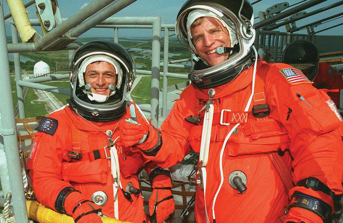 Due astronauti statunitensi in tuta arancione portano al polso un orologio digitale G-SHOCK nero.