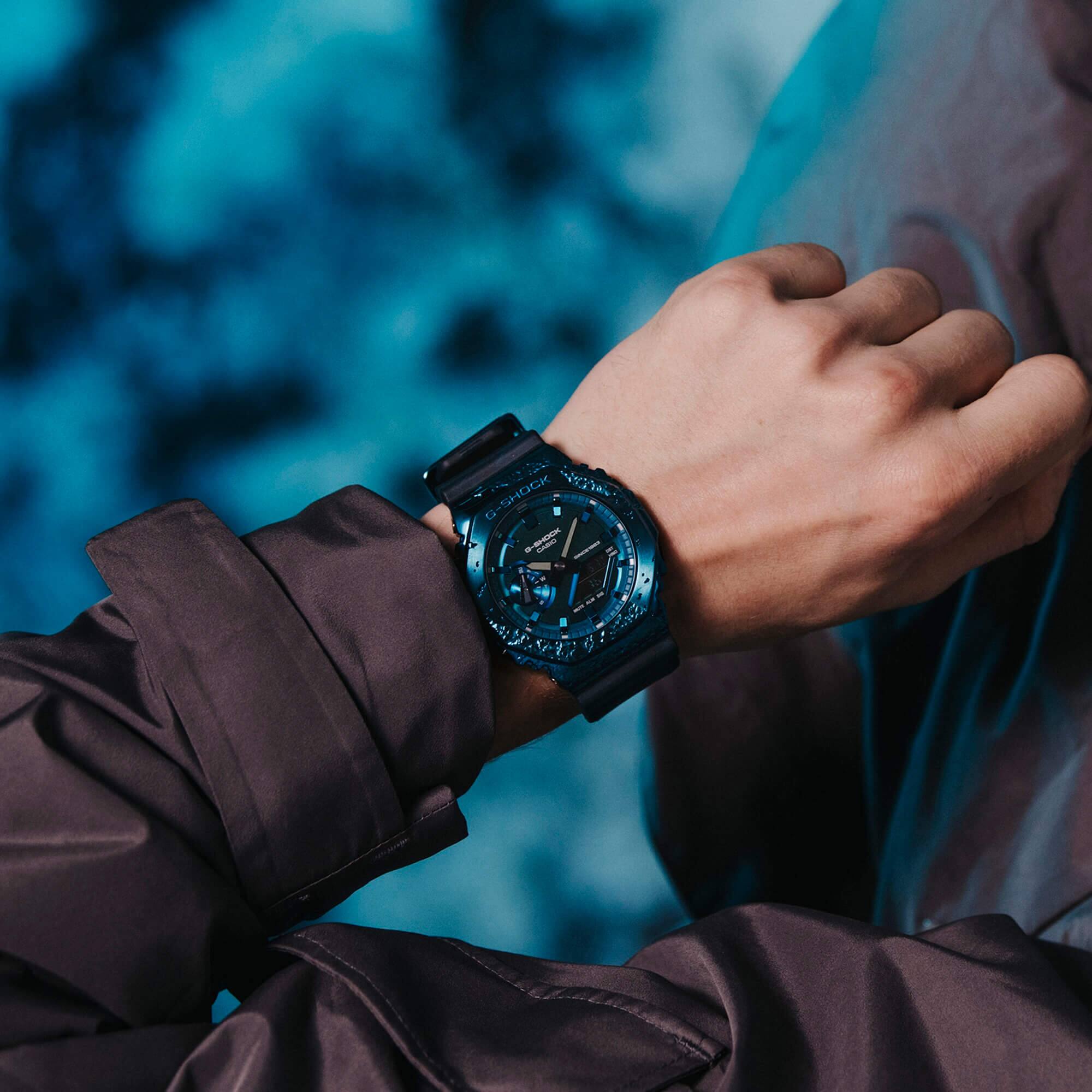 Vista dall'alto dell'orologio da polso G-SHOCK GM-2140GEM-2A con cassa turchese scintillante, display analogico e cinturino in resina nera al polso di un uomo.