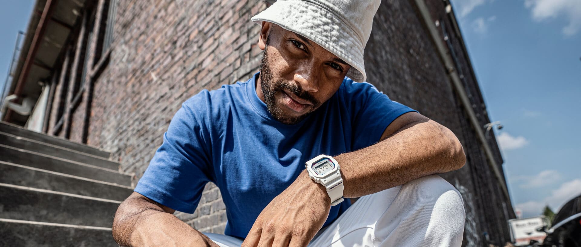 Un uomo guarda la telecamera con una camicia blu, un cappello da pesca bianco e un orologio digitale bianco della collezione Origin di G-SHOCK al polso.
