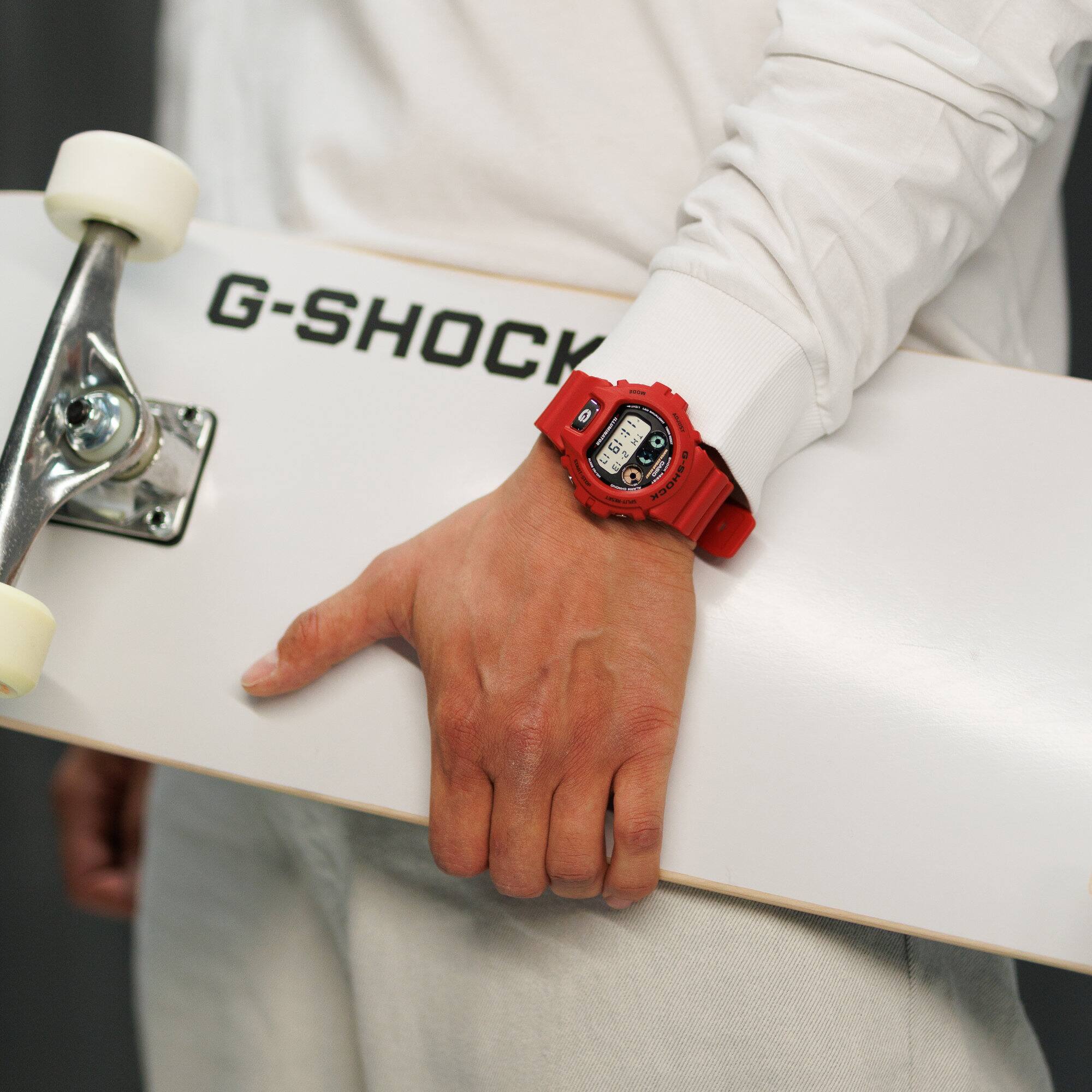 Un uomo vestito di bianco con uno skateboard e l'orologio digitale rosso “DW-6900TR-4ER” al polso.