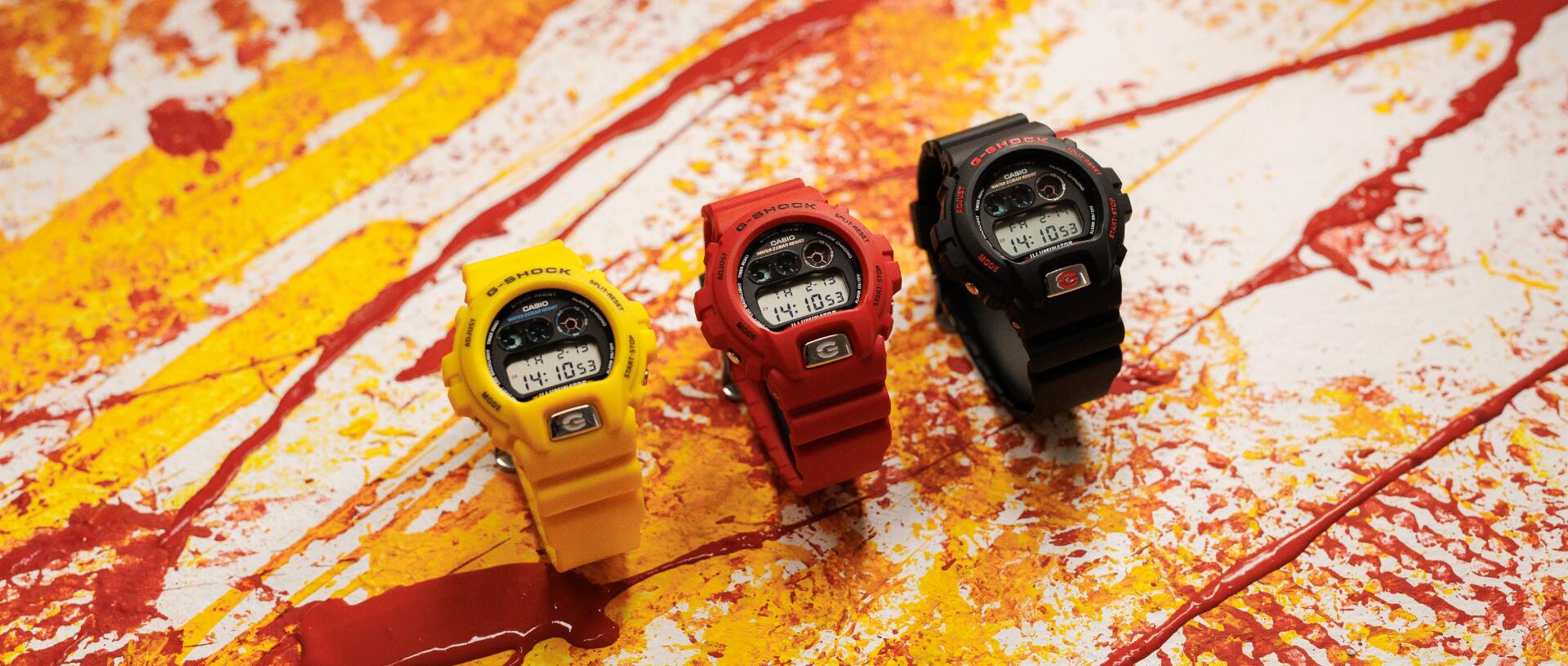 Tre modelli di orologio digitale DW-6900TR in giallo, rosso e nero su un terreno colorato a pioggia