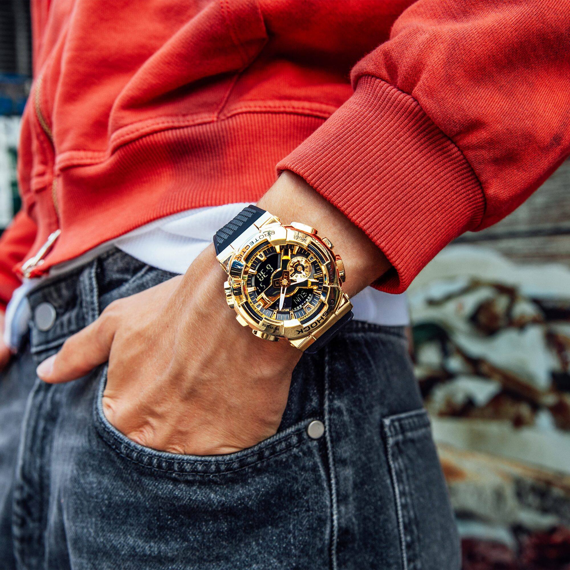 Primo piano di una persona in jeans e pullover rosso che indossa il GM-110 con custodia dorata e fascia nera al polso