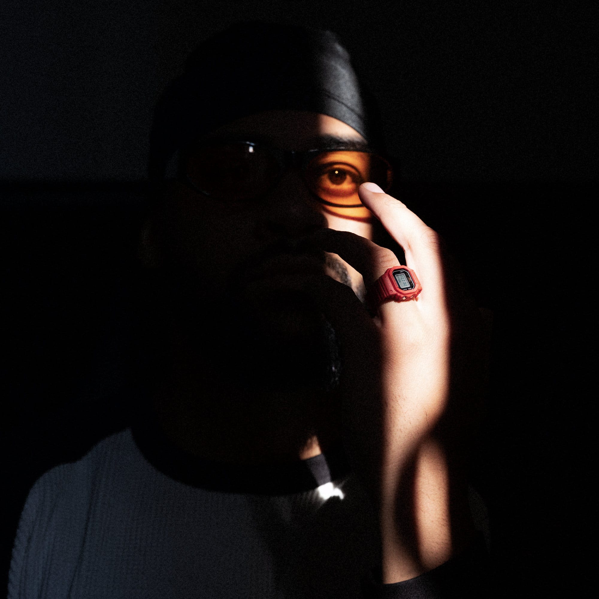 Primo piano sul volto di un uomo con occhiali arancioni, in penombra, che alza la mano. Indossa l'orologio digitale ad anello rosso DWN-6900 di G-SHOCK.