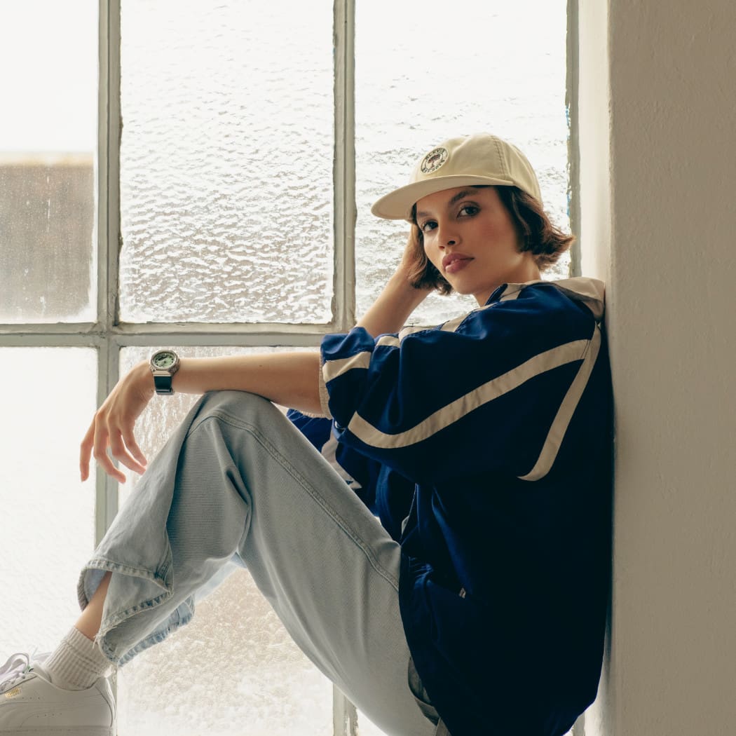 Una donna bruna con un berretto siede nella cornice di una finestra, appoggiando il braccio sul ginocchio e indossando il G-SHOCK "GM-S2110-3AER" al polso.