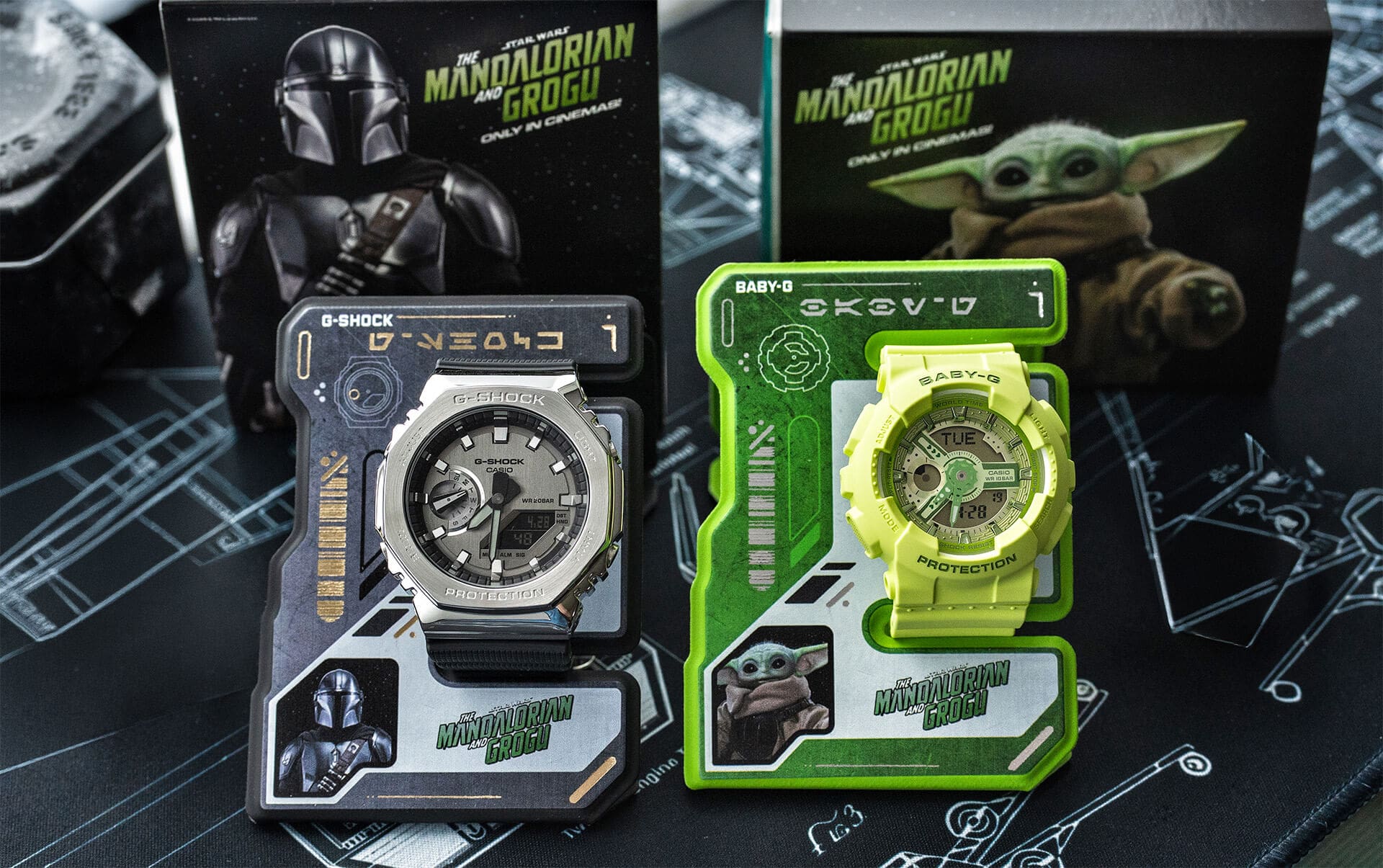 Twee horloges op de horlogestandaard met het Mandalorian- en Grogu-logo: de G-SHOCK GM-2100 met een metalen lunette en een analoog/digitaal display, en de BA-110 met een groen, rond analoog/digitaal display.