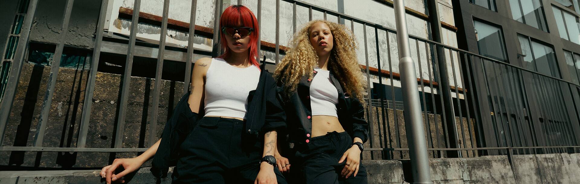 Een jonge vrouw leunt tegen een reling met een zwart jasje, een zwarte broek, een wit shirt en het zwarte GMA-P2100BB-1AER horloge van G-SHOCK om haar pols.