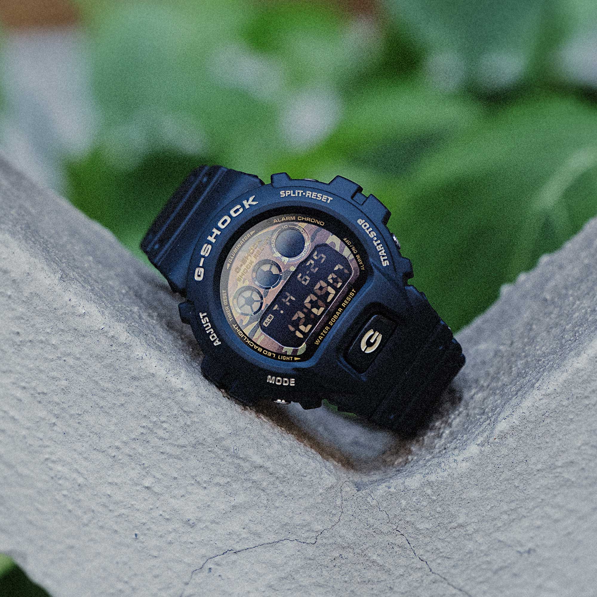 Het zwarte G-SHOCK DW-6900CF-1 digitale horloge met goudkleurige wijzerplaat ligt schuin op een lichte tak tegen een onscherpe groene achtergrond.