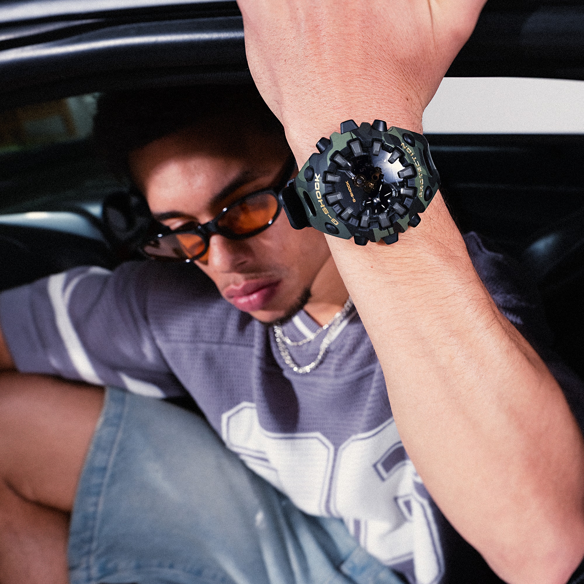 Een man met een zonnebril en een paars shirt stapt uit een auto en houdt zich vast aan het montuur. Het grote GA-V01-horloge is duidelijk zichtbaar om zijn pols.