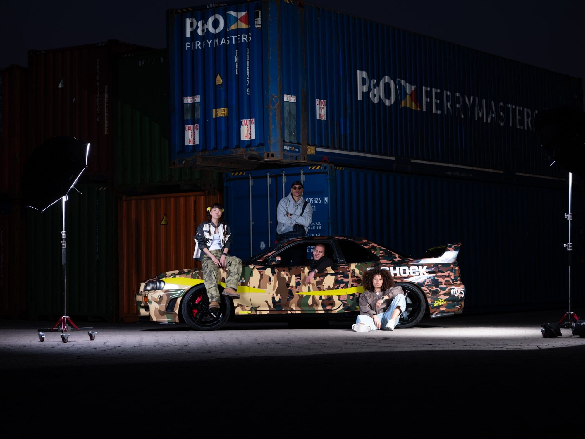 Een groep van vier zit of hurkt voor de Nissan, die geparkeerd staat in de haven voor containers.
