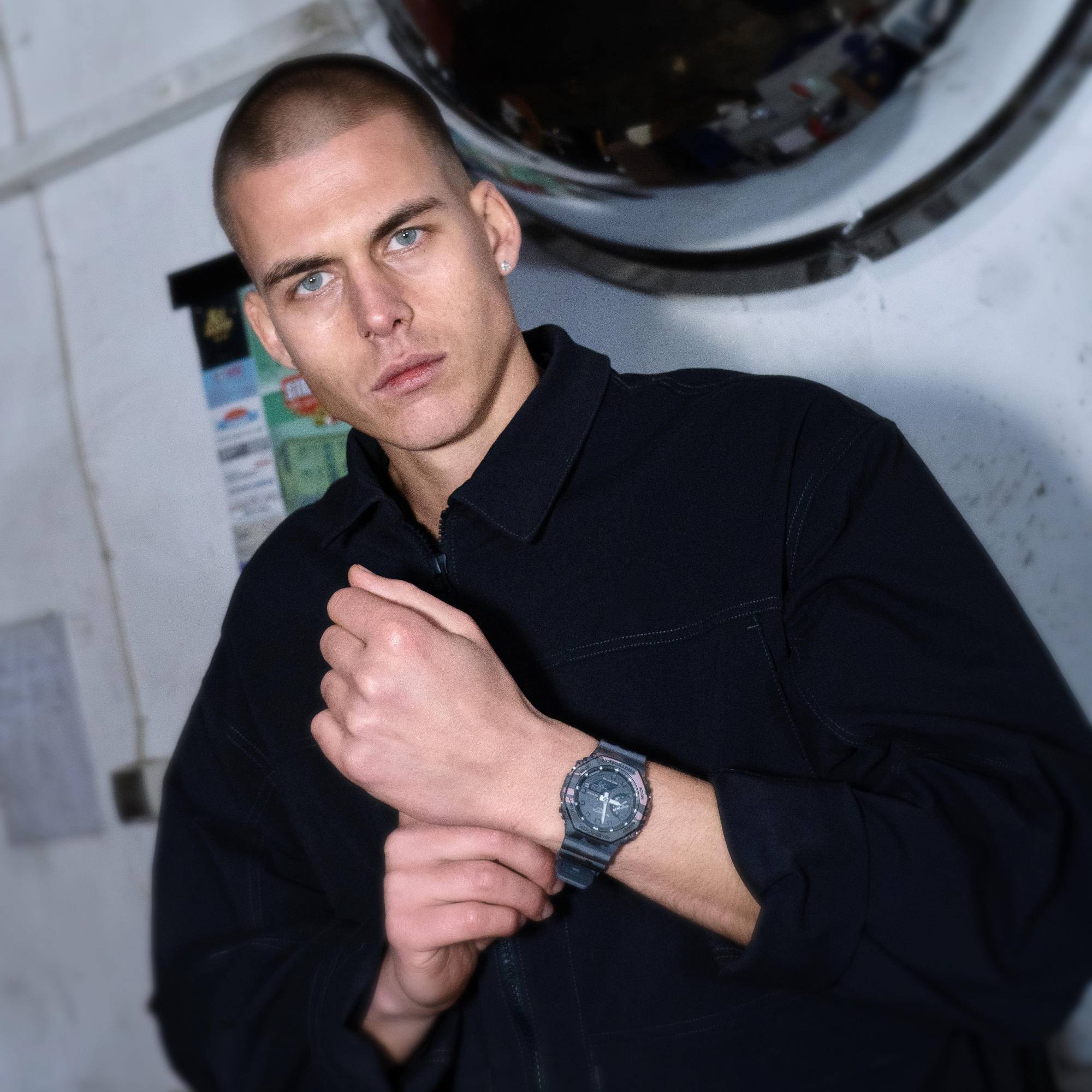 Een man in donkere kleding staat in een werkplaats en draagt een G-SHOCK GA-2100CM-8AER om zijn pols.