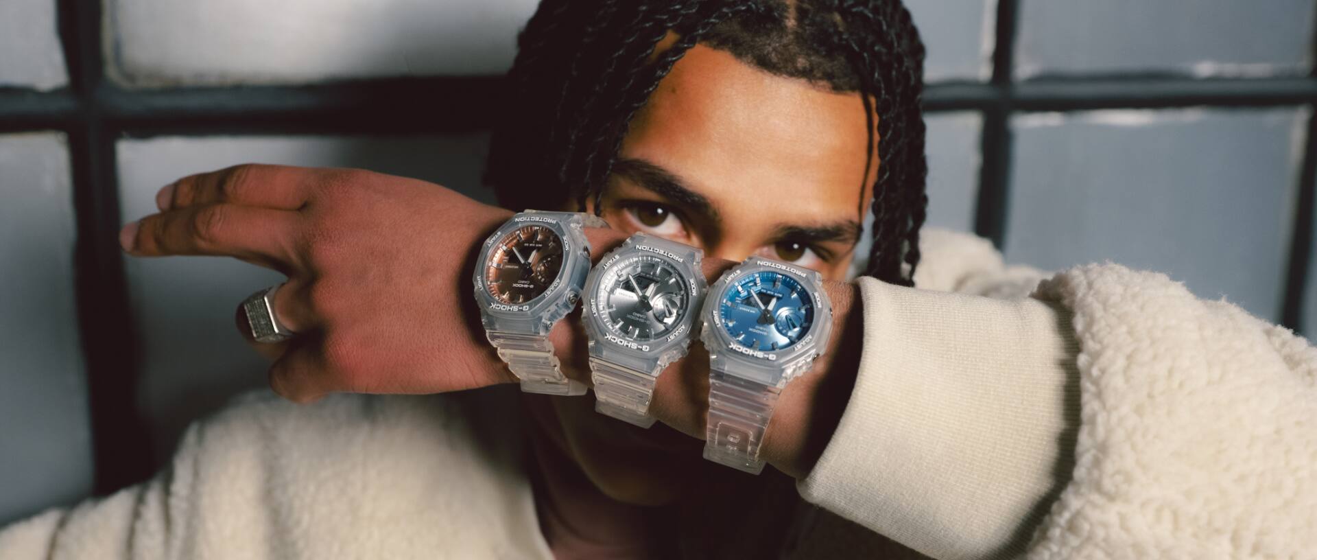 Drie G-SHOCK horloges met transparante armbanden en glinsterende lunetten om de pols van een man die de horloges voor zijn gezicht houdt.