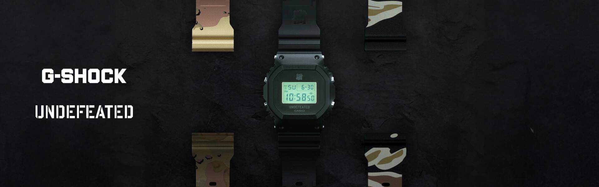 Het CASIO G-SHOCK DWE-5600UD-1 digitale horloge met lichtgevend LCD-display tegen een zwarte achtergrond. Links van het horloge met zijn transparante zwarte band is de vervangingsband in stormcamouflagedessin, rechts is de vervangingsband in tijgercamouflagedessin