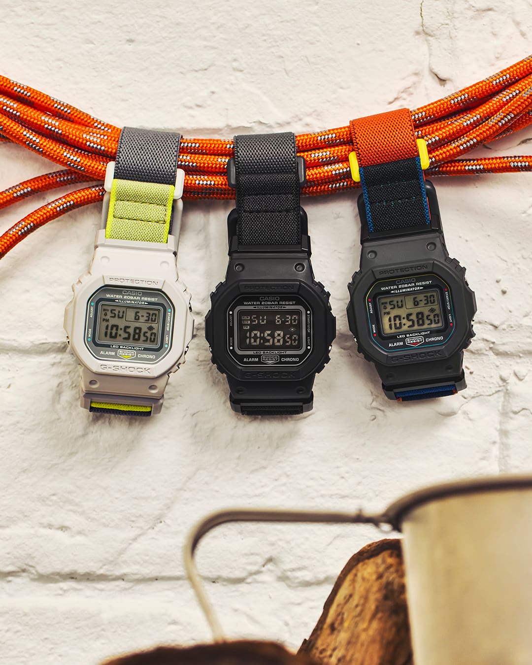 Drie G-SHOCK DW-5600MNC-modellen in verschillende kleurstellingen, opgehangen aan meerdere touwen voor een gestructureerde muur.