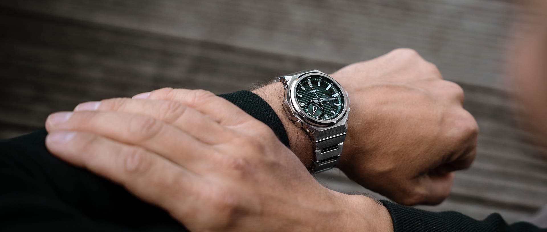 Het G-SHOCK G-Steel horloge GST-B1000 met een zilverkleurige band, een donkergroene analoge lunette en witte indexen in de pols van een man