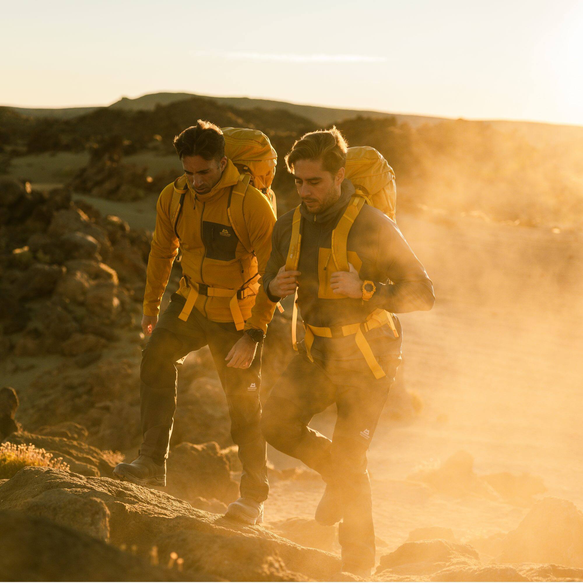 Twee mannen lopen buiten op stoffige grond in de avondzon en dragen rugzakken en de G-SHOCK Rangeman.