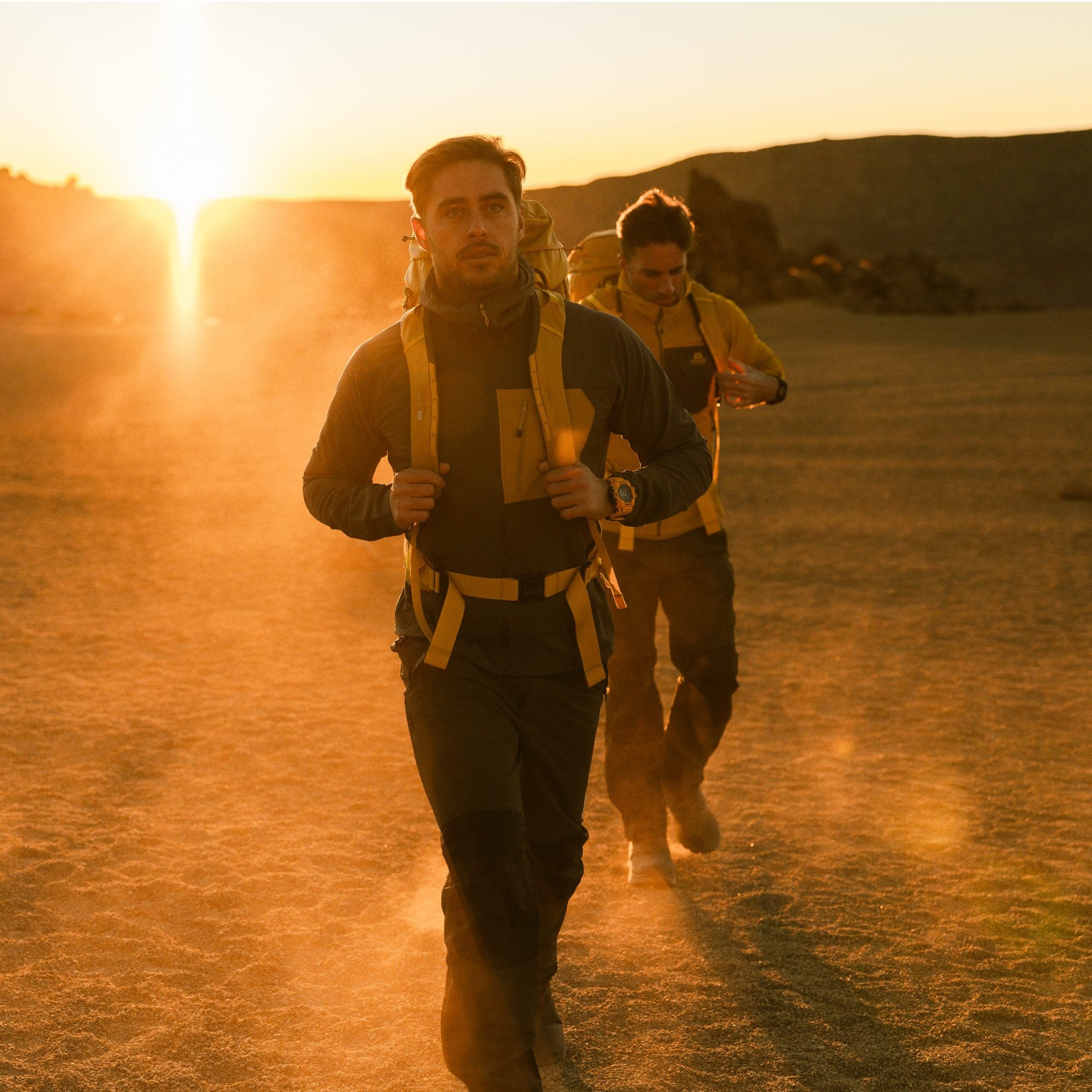 Twee mannen lopen buiten op stoffige grond in de avondzon en dragen rugzakken en de G-SHOCK Rangeman.