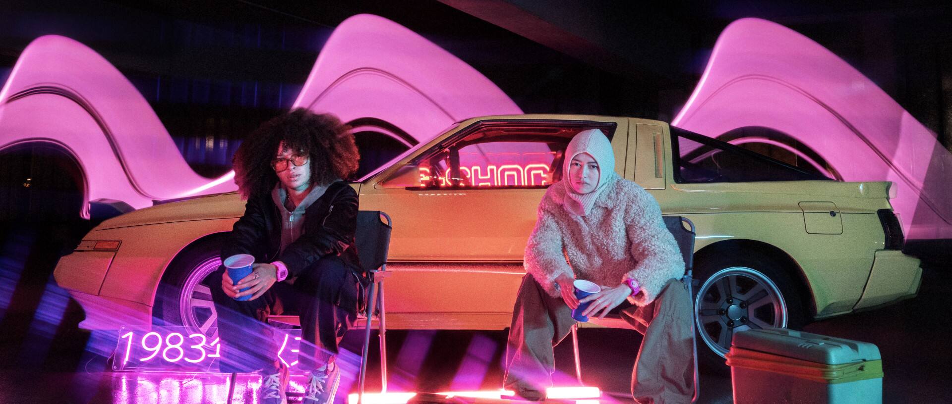Twee vrouwen zitten in een donkere parkeergarage voor een gele auto naast neonroze lichten met roze G-SHOCK horloges om hun pols.