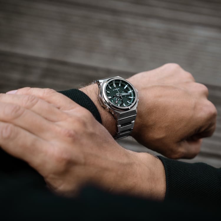 Vista aérea do pulso de um homem que usa o relógio GST-B1000 da G-SHOCK com bracelete prateada e mostrador redondo verde escuro. O link leva à página inicial dos relógios G-STEEL GST-B1000.