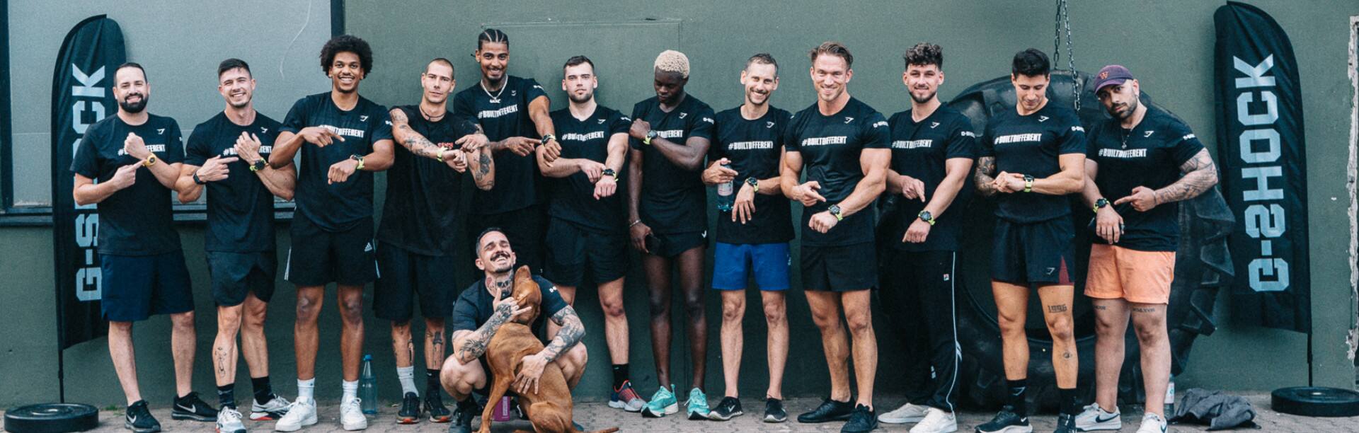 Um grupo de homens em trajes desportivos curtos pretos em frente a um muro de betão, todos apontando para o pulso no qual usam o G-Squad GBD-H2000