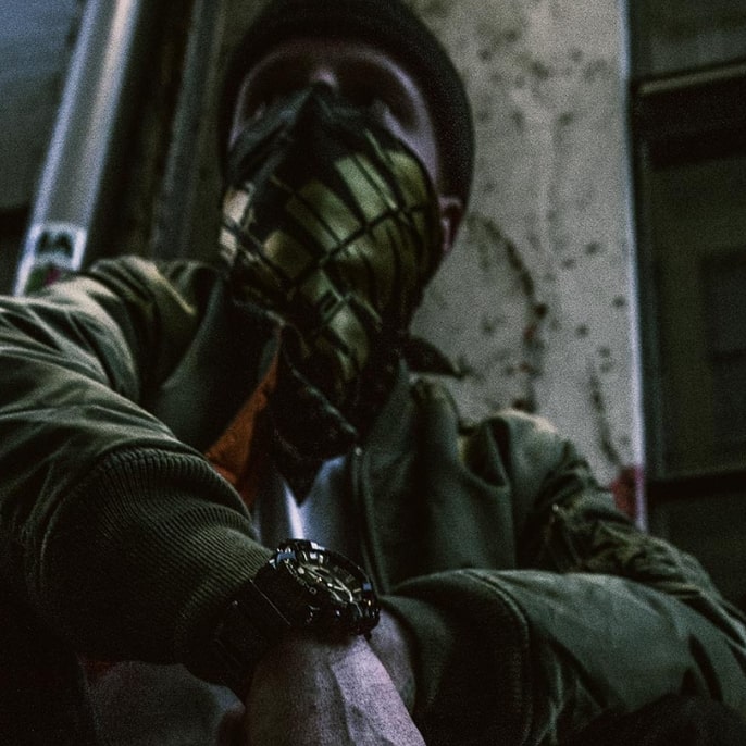 Uma pessoa com um lenço à frente do rosto agacha-se contra uma parede e usa o relógio G-SHOCK no pulso
