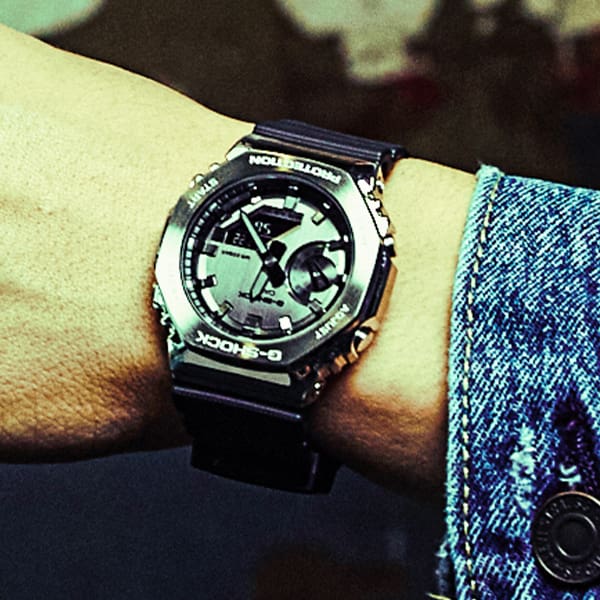 O relógio de pulso analógico GM-2100-1A com bracelete de resina preta e caixa octogonal de metal no pulso de uma pessoa com um casaco de ganga.