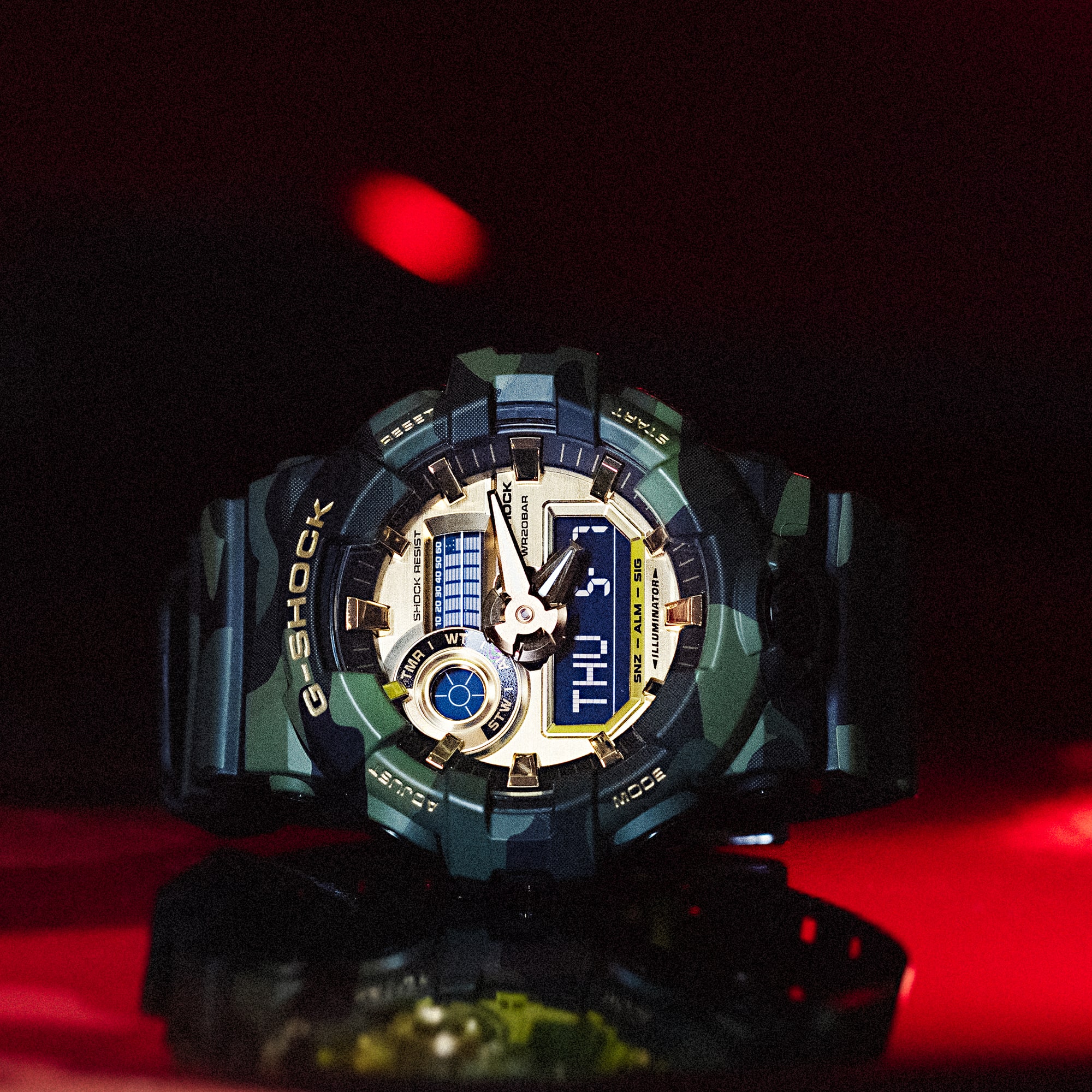 O GA-700CMG-3A, com seu mostrador analógico-digital, está deitado de lado contra um fundo vermelho. O visor brilha em tons dourados e a pulseira de resina apresenta um padrão camuflado.
