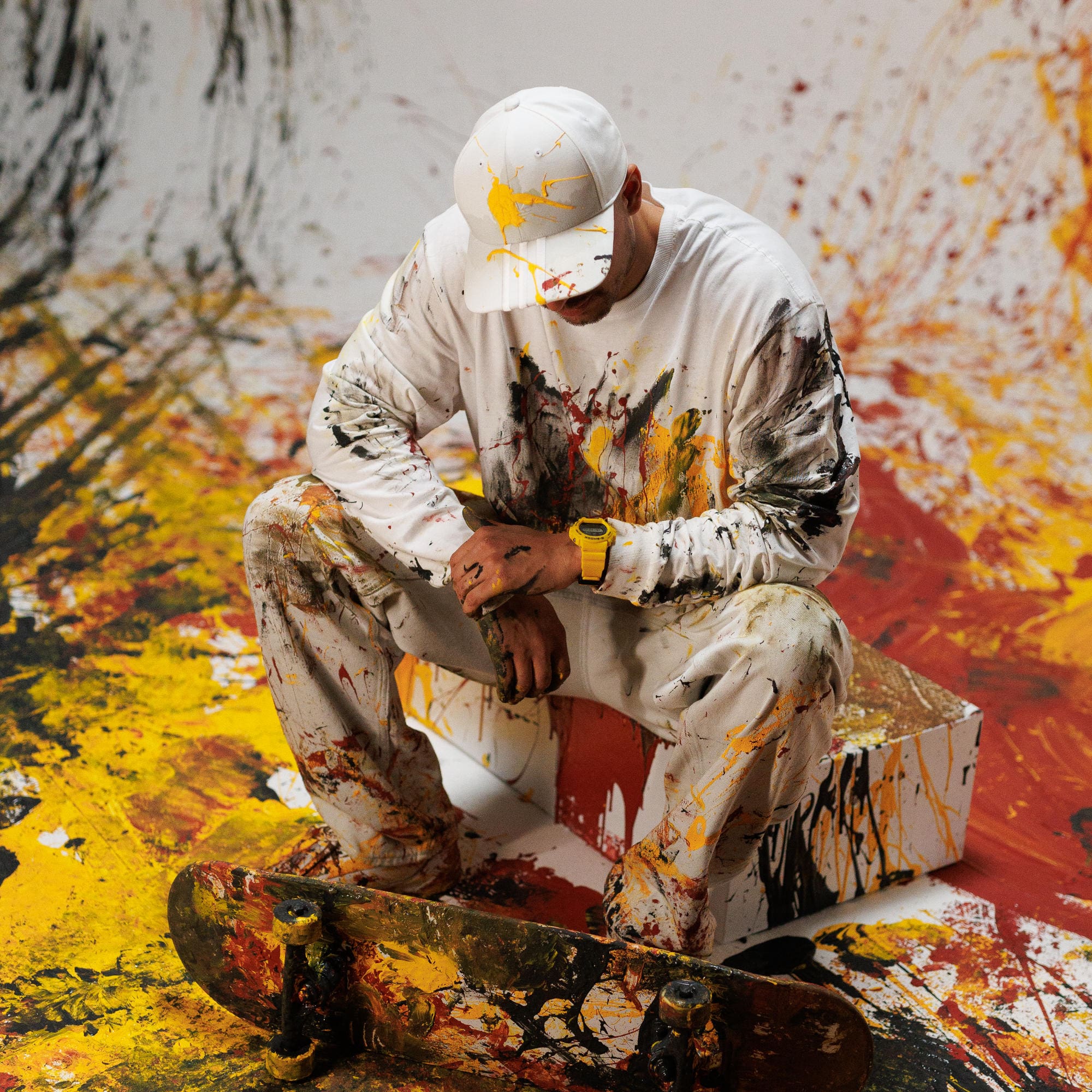 Um homem sentado numa rampa ao lado do seu skate numa sala salpicada de vermelho, preto e amarelo