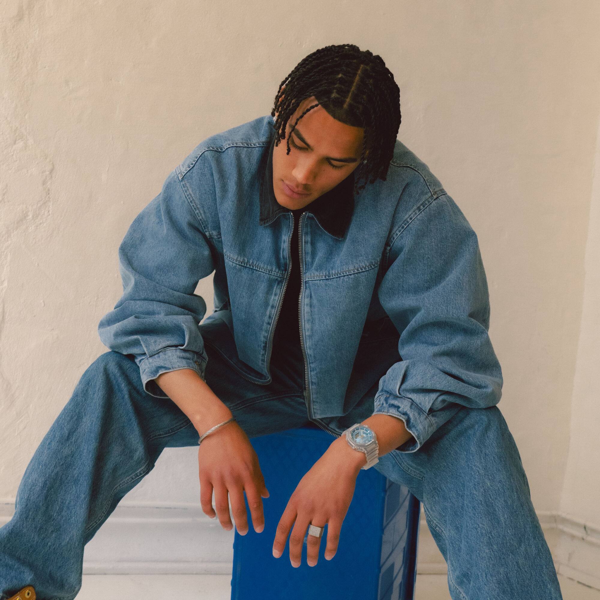 Um homem de calças de ganga senta-se num banco azul e inclina-se para a frente, usando um G-SHOCK GA-2100BM com um mostrador azul claro no pulso.