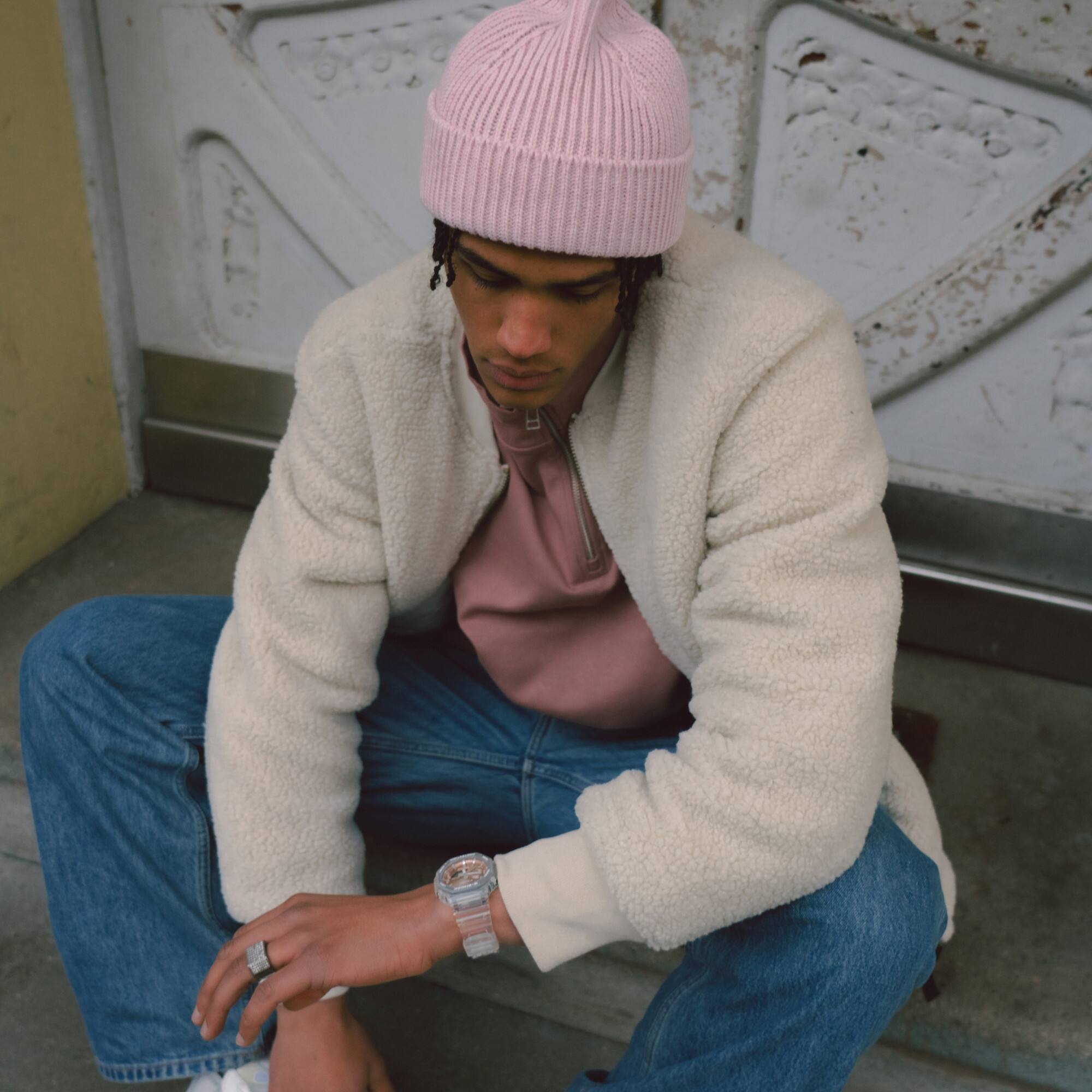 Um homem com uma camisa cor-de-rosa, boné cor-de-rosa e calças de ganga senta-se num degrau e olha para o seu pulso, no qual está a usar o GA-2100BM com uma luneta cor-de-rosa e laranja.