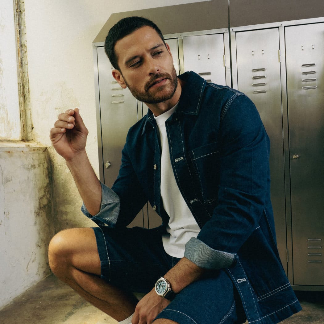 Um homem de calças de ganga senta-se num banco, olha para a direita e usa o relógio de metal G-SHOCK "GBM-2100" no pulso