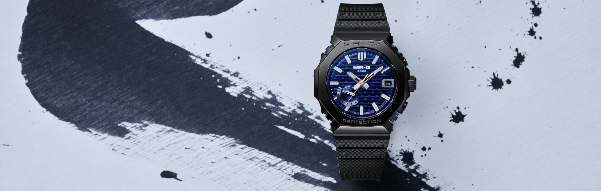 O MRG-B2100R com bracelete preta e luneta azul meia-noite sobre um fundo pingado de tinta.