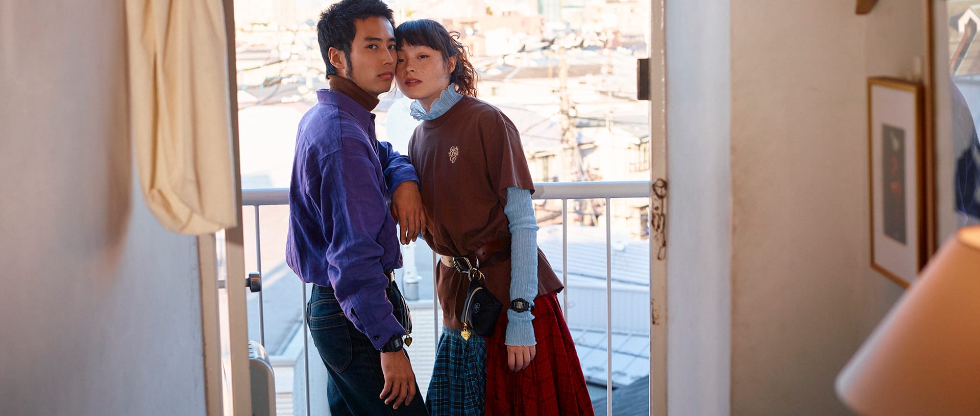 G-SHOCK x VERDY (Girls Don't Cry) collaboration. A male and female model standing close together on a balcony, looking at the camera. The man wears a purple shirt, the woman a brown Girls Don't Cry logo t-shirt, each wearing a G-SHOCK watch on their wrist.