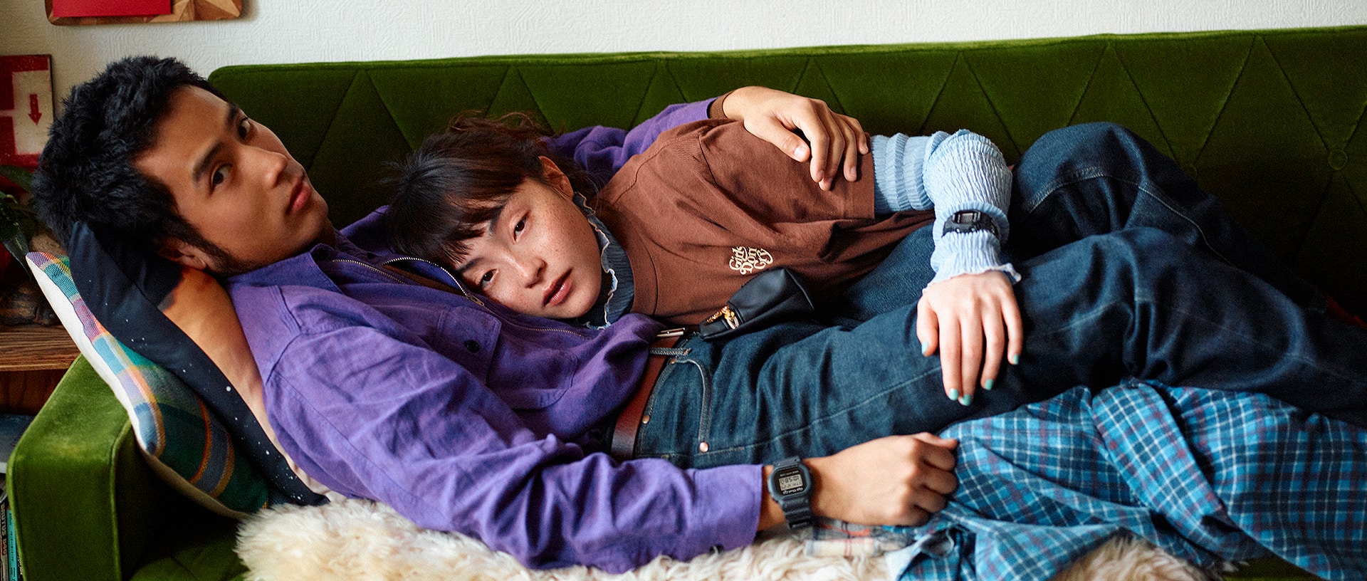 G-SHOCK x VERDY collaboration. A couple lying close together on a green sofa. The man wears a purple shirt, the woman a brown t-shirt with the Girls Don't Cry logo, and both have G-SHOCK watches on their wrists.