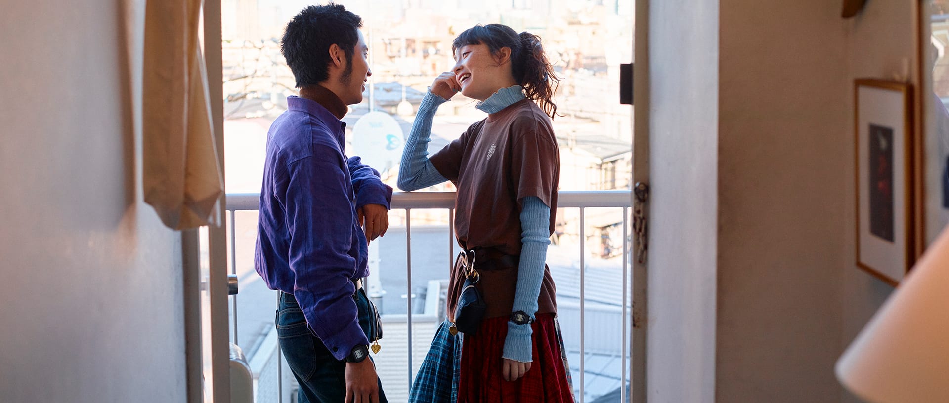G-SHOCK x VERDY (Girls Don't Cry) collaboration. A male and female model smiling and facing each other on a sunlit balcony. The man wears a purple shirt, the woman a brown t-shirt layered with arm covers, and wears a G-SHOCK watch over the arm cover.