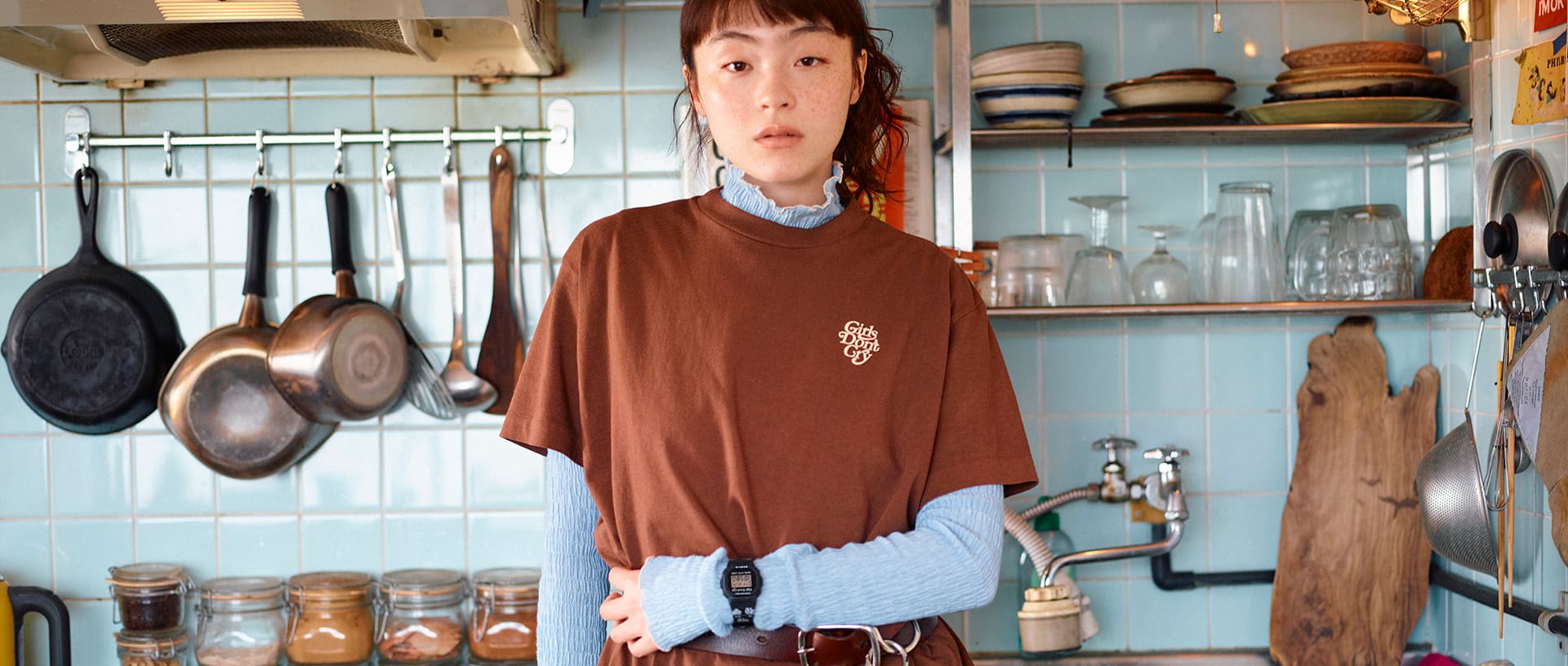 G-SHOCK x VERDY (Girls Don't Cry) collaboration. A female model standing in a retro kitchen, wearing a brown t-shirt with the Girls Don't Cry heart logo on the chest and a G-SHOCK watch over her sleeve.