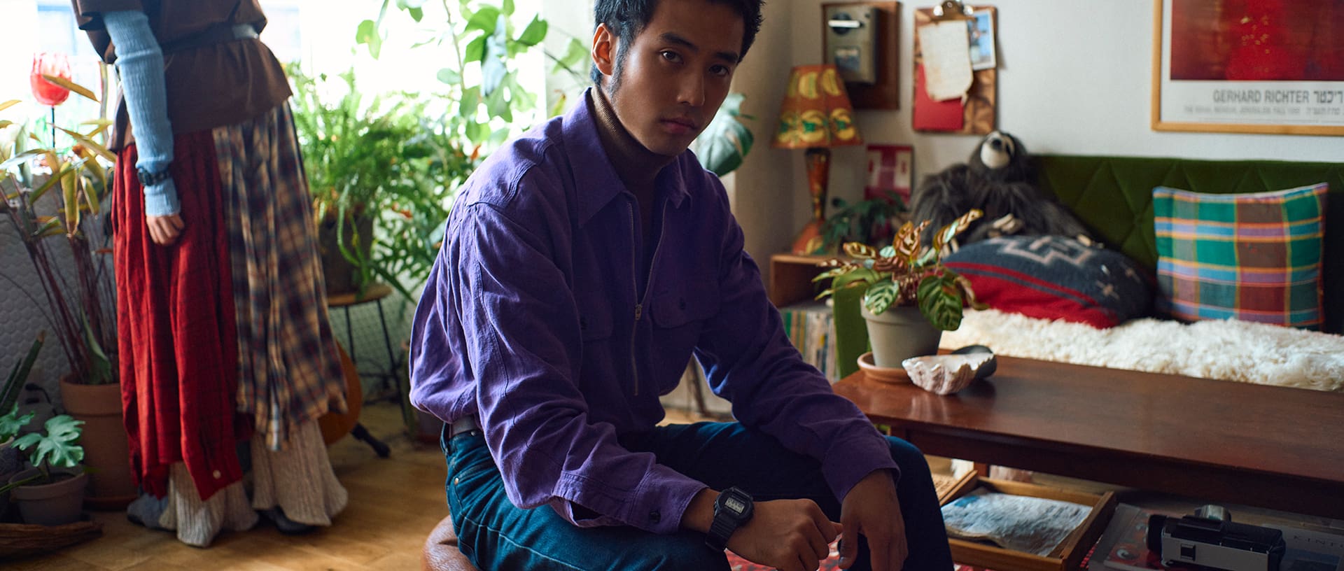G-SHOCK x VERDY (Girls Don't Cry) collaboration. A male model sitting in a room with houseplants and a sofa, highlighting the G-SHOCK watch on his wrist, with a female model standing in the background.