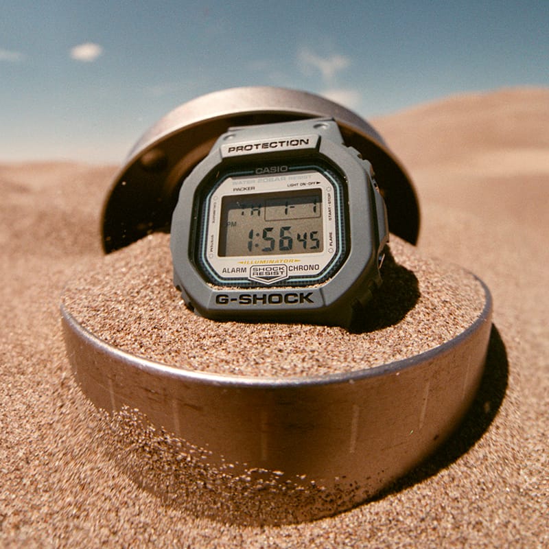 G-SHOCK X PACKER DW5600PCK23-2B gray digital watch in a sand dune