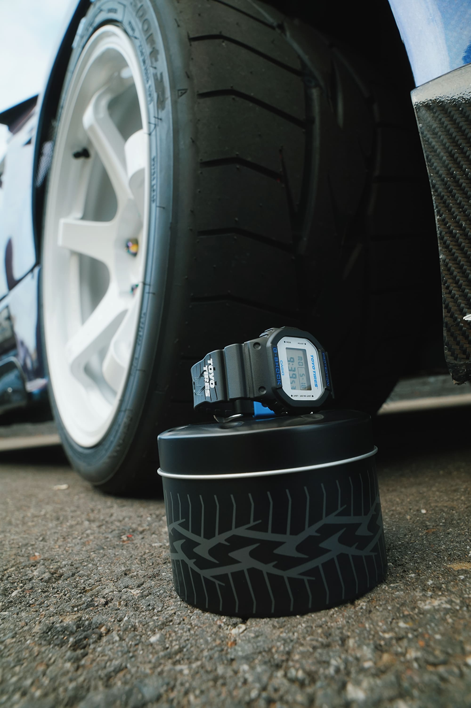 G-SHOCK x TOYO TIRES DW5600TT25-1 laid on special tin packaing beside the Proxes Sport R tire that inspired the watch design