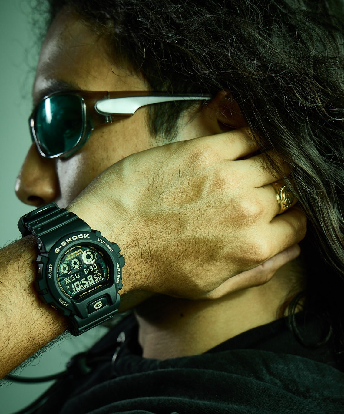 Long-haired person wearing sunglasses and a black digital G-SHOCK watch with one hand near their face.