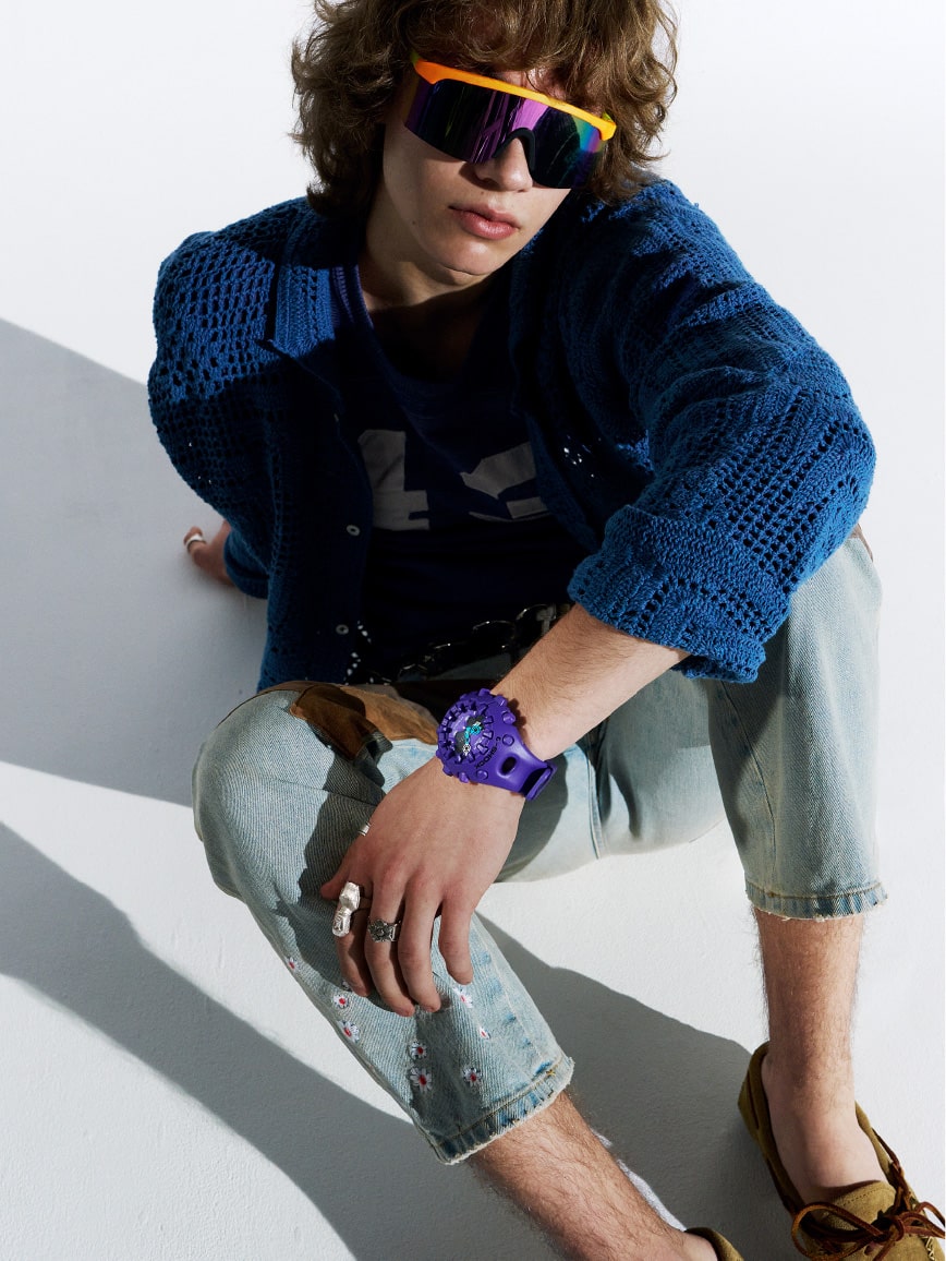شخص يجلس بارتياح ويرتدي ساعة G-SHOCK باللون الأرجواني ونظارات شمسية.
