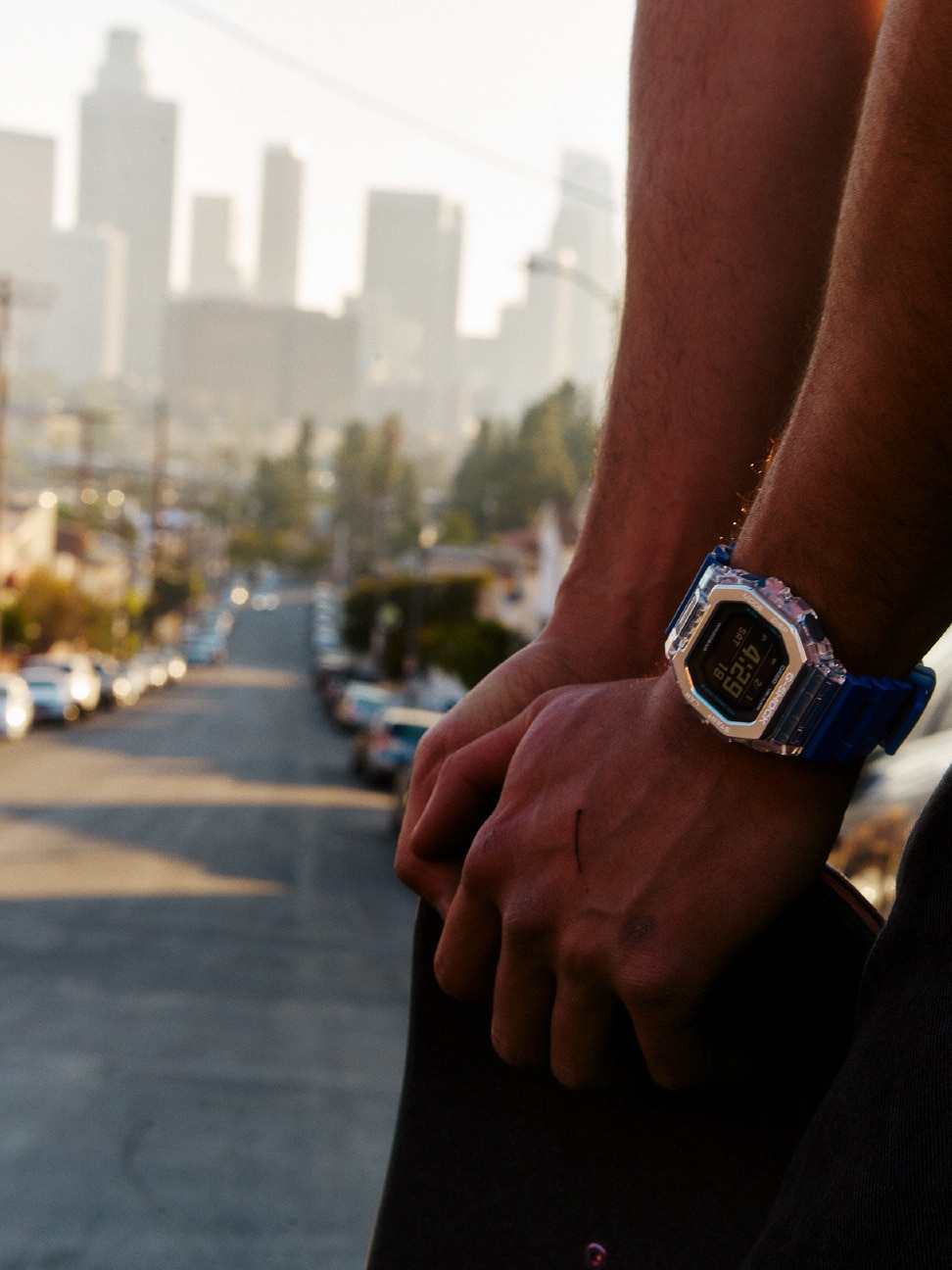 شخص يرتدي ساعة G-SHOCK باللون الأزرق ويحمل لوح تزلج، في موقع يطل على المدينة.