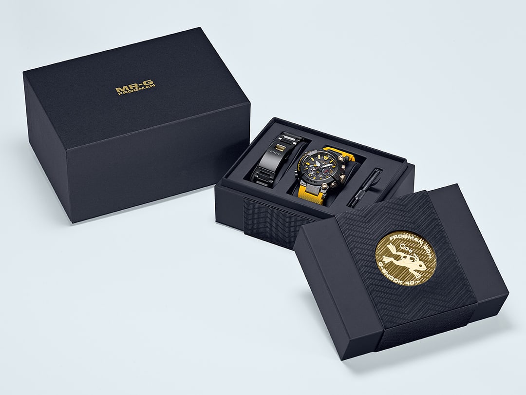Der dunkle Umkarton mit goldener MR-G Frogman-Prägung neben der offenen Box mit der MRG-BF1000E-1A9 Armbanduhr mit gelben Armband, außerdem in der Box: das Edelstahlarmband und Montagewerkzeug